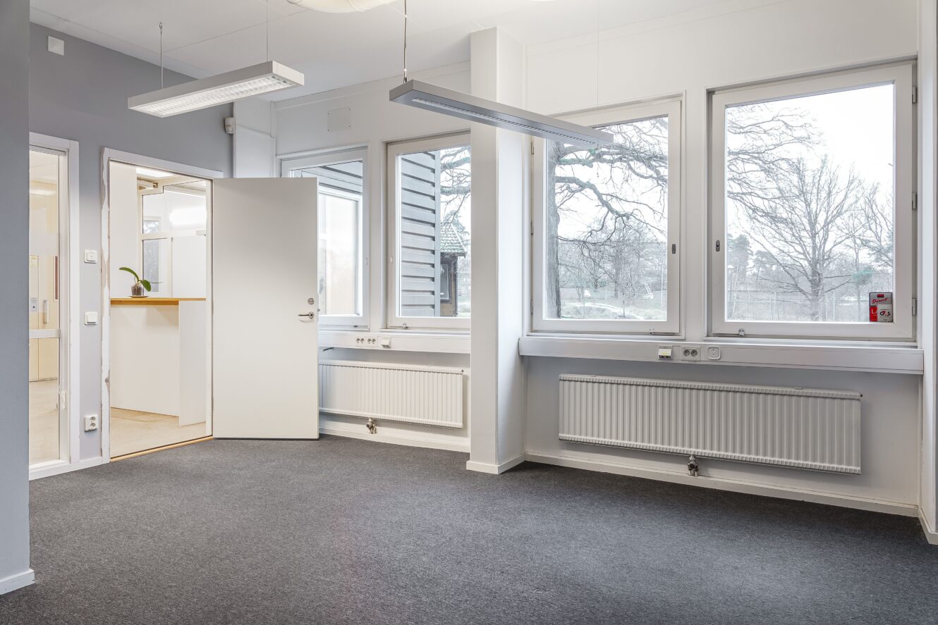 Ett tomt kontorsrum med grå heltäckningsmatta, vita väggar, stora fönster, element och en dörr som leder ut till en hall.