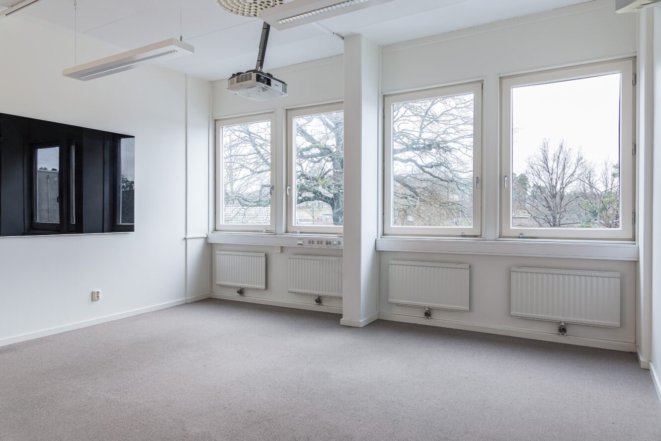 Tomt kontorsrum med stora fönster, väggmonterad skärm, projektor, vita väggar, element och neutral heltäckningsmatta. Träd syns utanför fönstren.