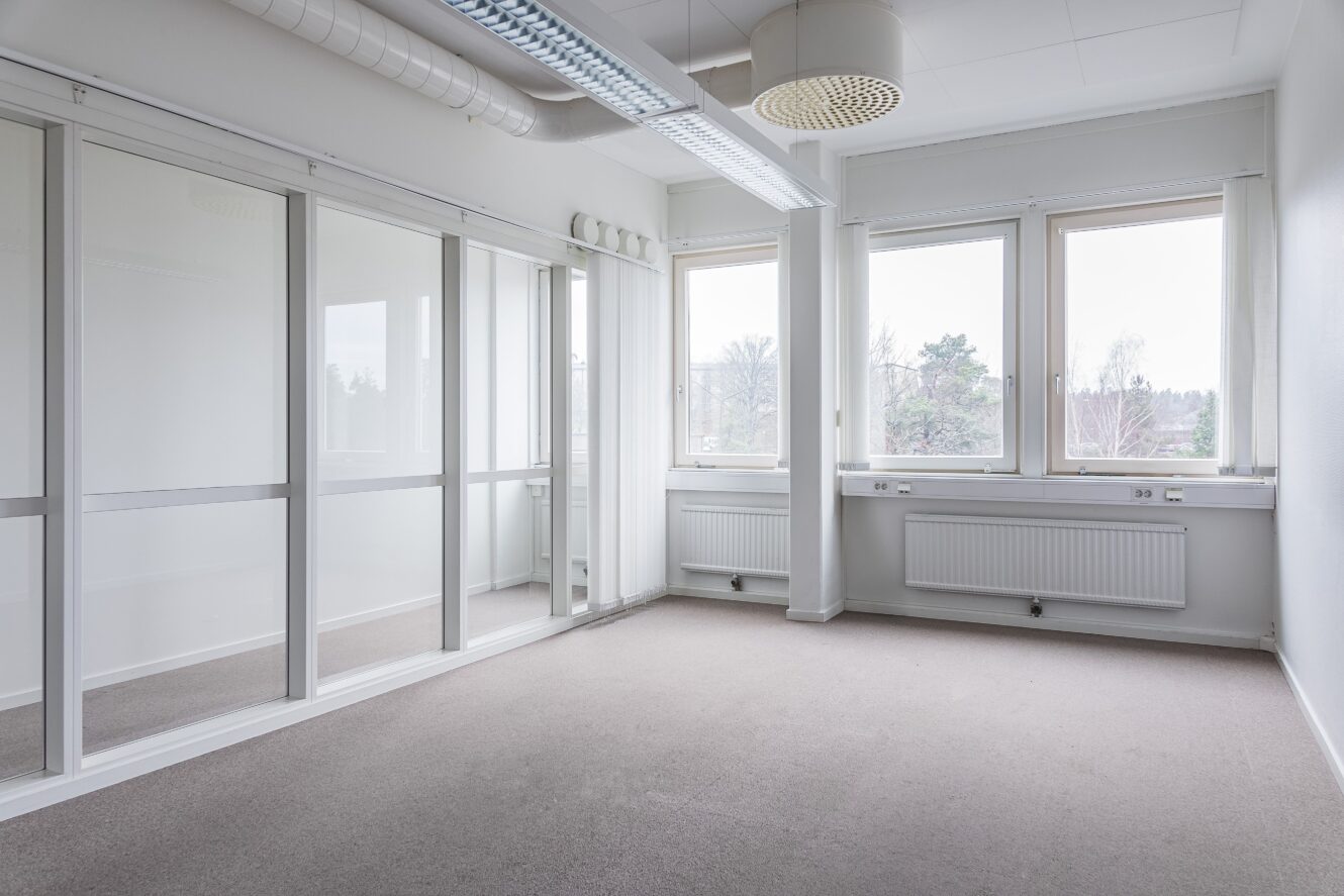 Ett tomt, omöblerat kontorsrum med stora fönster, glasväggar, vita väggar och beige matta. Naturligt ljus kommer in genom fönstren som vetter utåt.