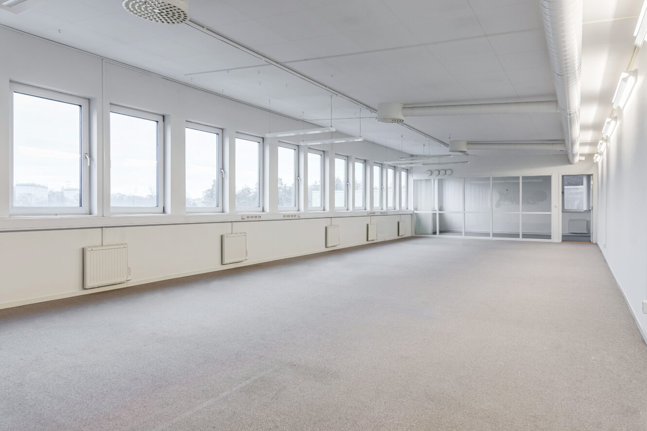 Tomt kontorsutrymme med stora fönster längs ena väggen, vita väggar, beige heltäckningsmatta, taklampor och glasvägg längst bort.