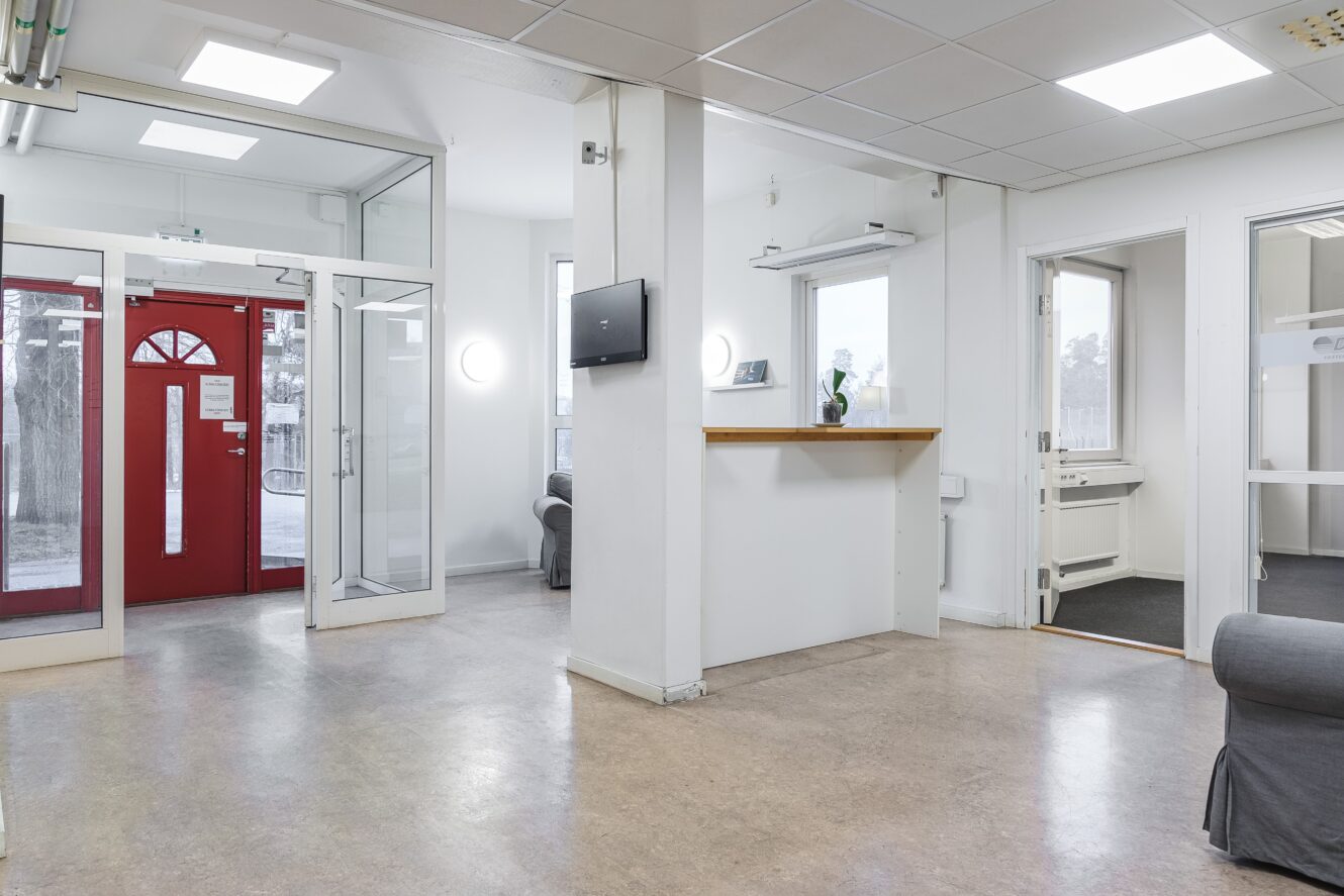 Ljus och stilren lobby med vit receptionsdisk, röda dubbeldörrar, glaspaneler och gråa sittmöbler. Lysrör och minimalistisk inredning skapar en modern och funktionell atmosfär.