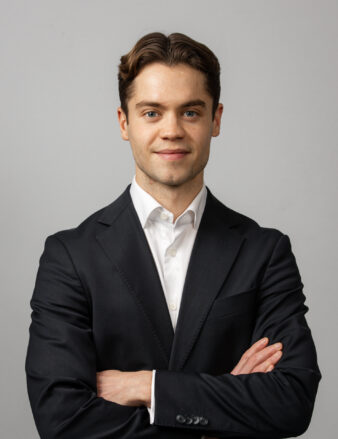 En ung man i mörk kostym och vit skjorta står mot en grå bakgrund med armarna i kors och tittar rakt in i kameran.