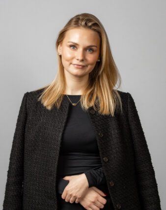 En kvinna med rakt blont hår, klädd i en svart texturerad jacka över en svart topp, står framför en vanlig ljusgrå bakgrund.