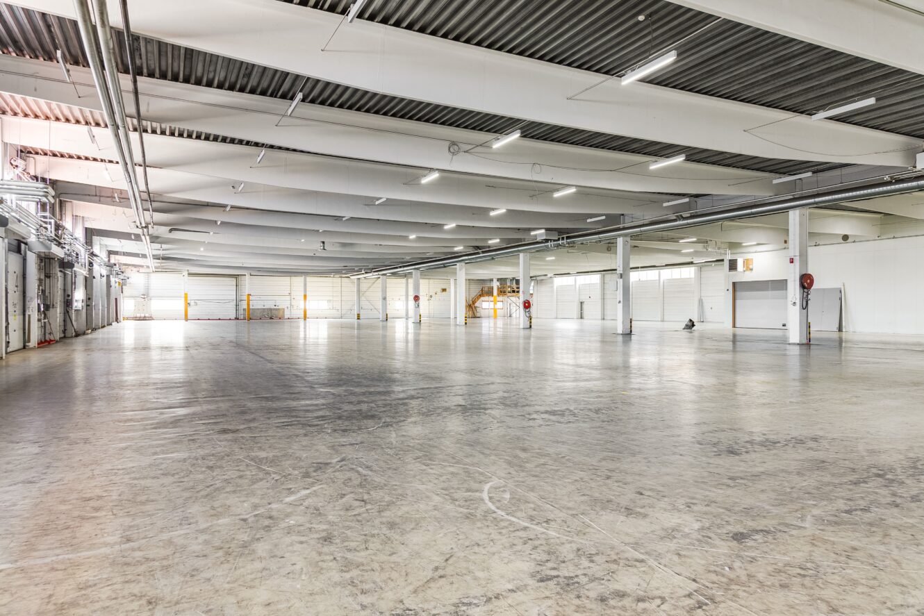 Stort, tomt industrilager med polerade betonggolv, högt i tak, takbelysning och lastbryggor längs ena väggen.