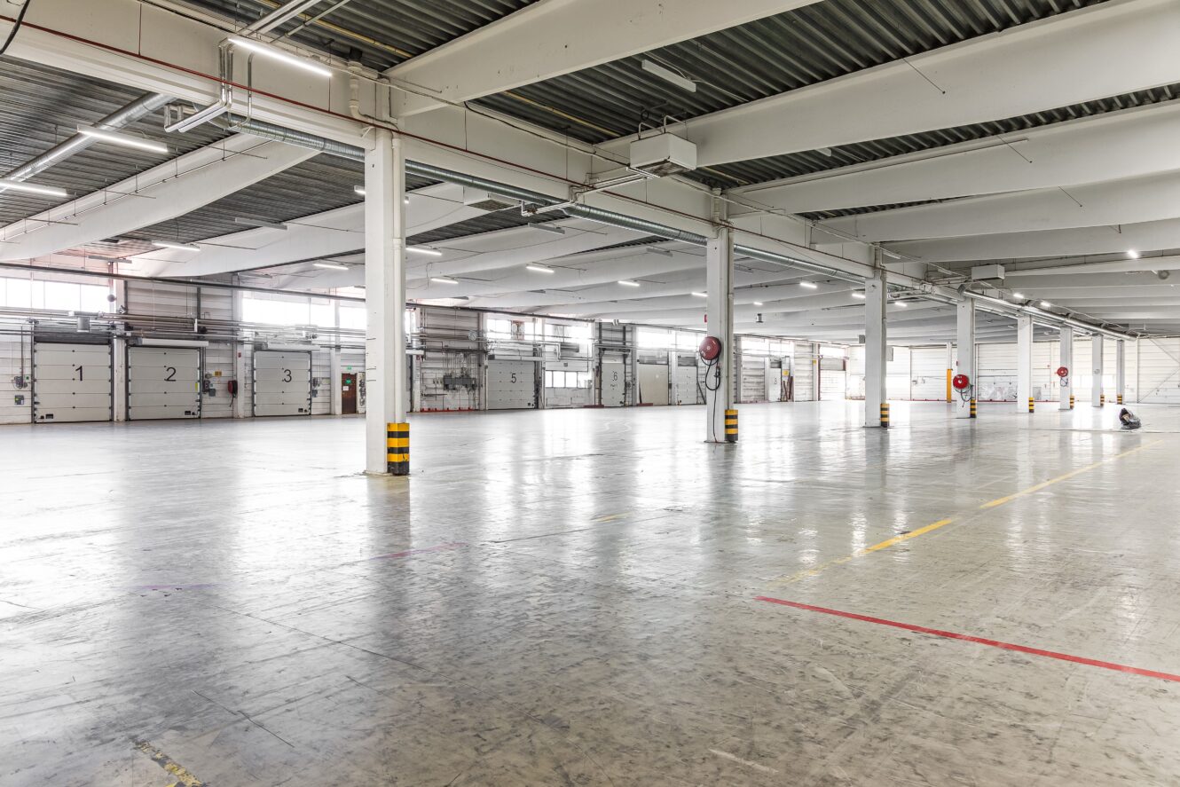 Stor, tom lagerlokal med betonggolv, stödpelare, högt i tak och flera lastkajsdörrar längs ena väggen.