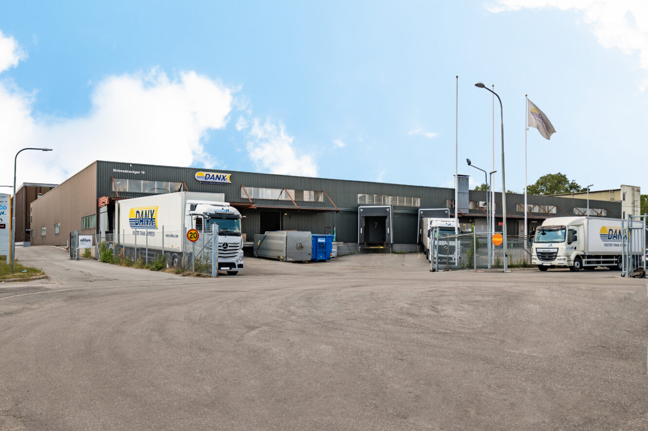 Industriell lagerlokal med DANX-skyltar, flera lastbilar parkerade vid lastbryggor och flaggor på stänger, under en delvis molnig himmel.