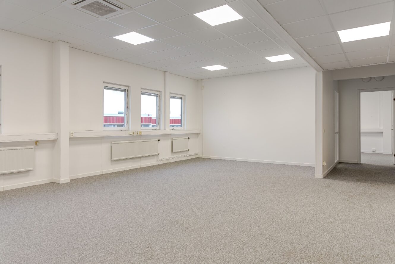 Tomt kontorsutrymme med vita väggar, grå heltäckningsmatta, tre fönster, taklampor och en öppen dörr som leder till ett annat rum.