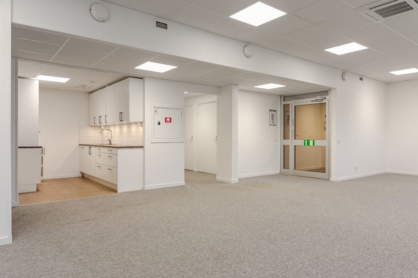 Omöblerat kontorsutrymme med pentry, vita väggar, heltäckningsmatta och lysrör i taket. Dubbla glasdörrar och nödutgångsskyltar är synliga.