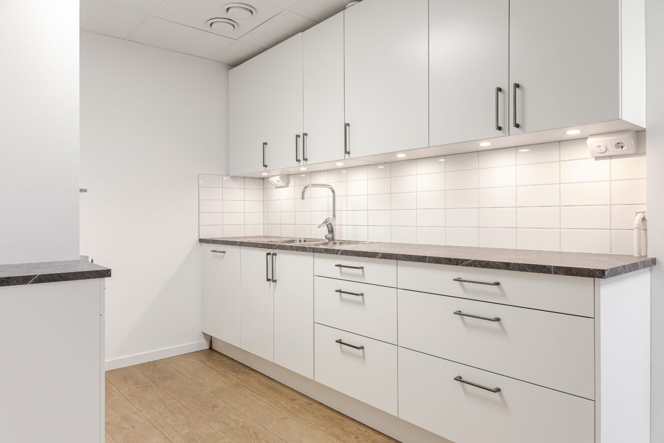 Modernt kök med vita skåp, belysning under skåpen, grå bänkskiva, kaklat stänkskydd och trägolv.