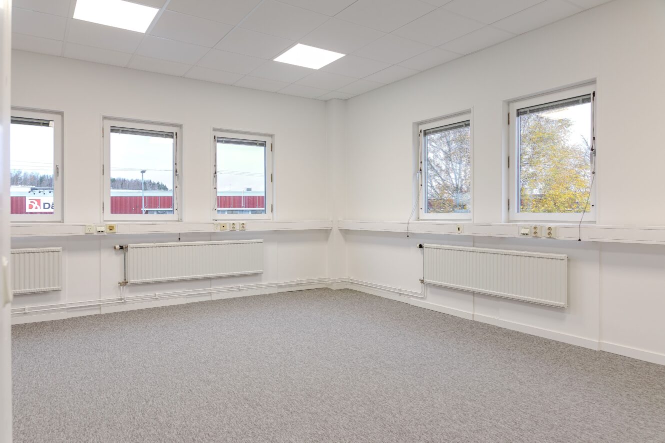 Tomt kontorsrum med grå heltäckningsmatta, vita väggar, tre fönster på ena sidan, två fönster på den andra och radiatorer under fönstren.