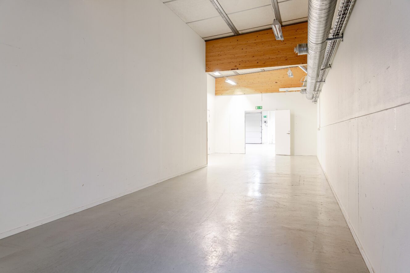 En tom, väl upplyst hall med vita väggar, ett polerat golv, synliga ventilationsrör och ett takparti i trä som leder till en öppen dörr med en nödutgångsskylt.