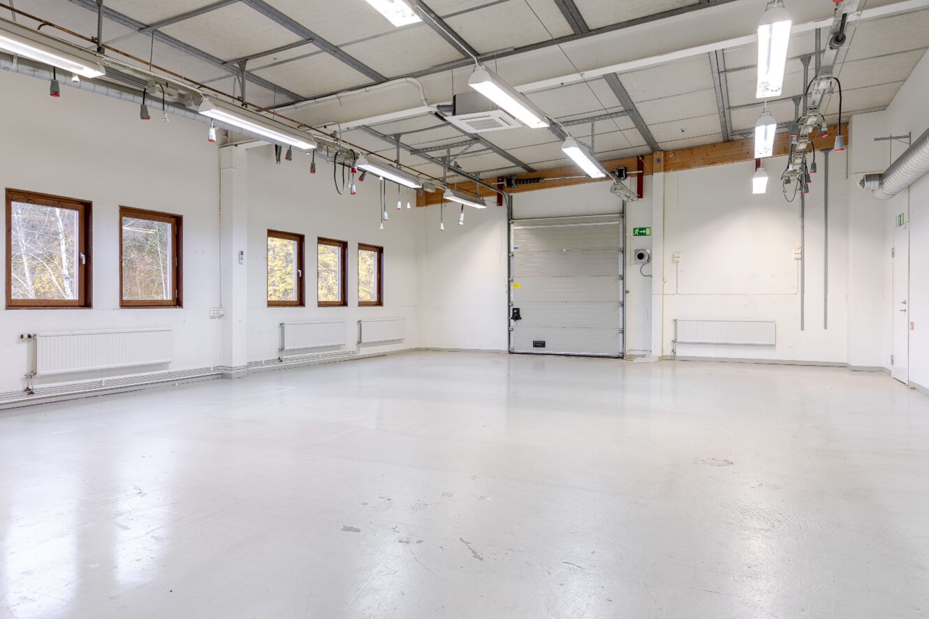 Tomt industrigarage med vita väggar, stora fönster, taklampor, väggmonterade element och en stängd rullport.