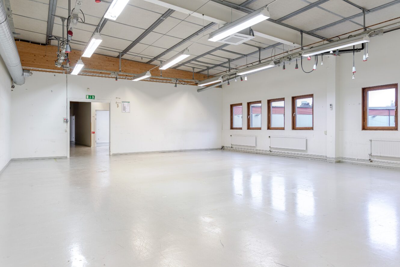 Tomt industrirum med vita väggar, stora fönster, synligt tak och lysrörsbelysning; synlig utgångsdörr och brandsäkerhetsutrustning på väggarna.