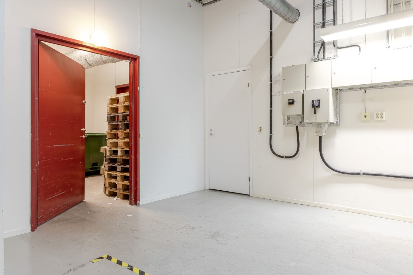 En ren tvättstuga med vita väggar, elpaneler på den högra väggen, en stängd vit dörr och en öppen röd dörr som avslöjar staplade träpallar och en grön soptunna.