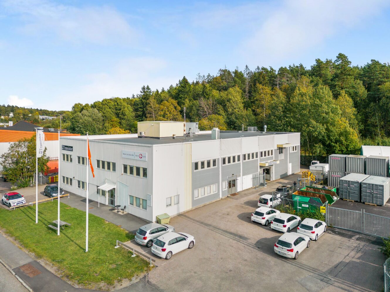 Flygfoto av en tvåvånings industribyggnad med flera parkerade bilar, flaggstänger, containrar och träd i bakgrunden.