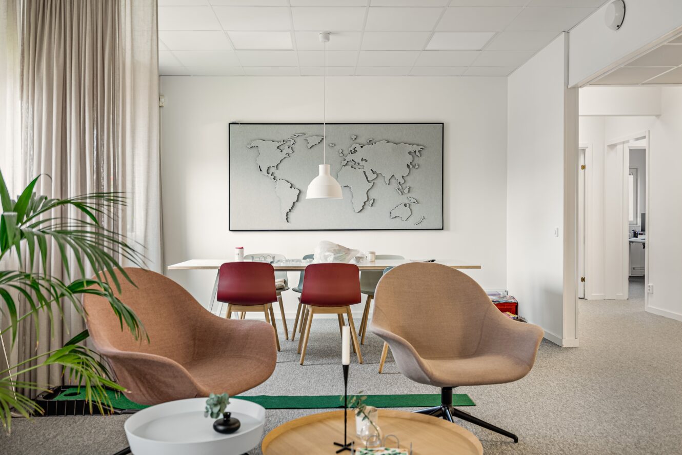 Modern kontorslounge med två bruna stolar, två röda stolar vid ett vitt bord, en världskarta på väggen och inomhusväxter.