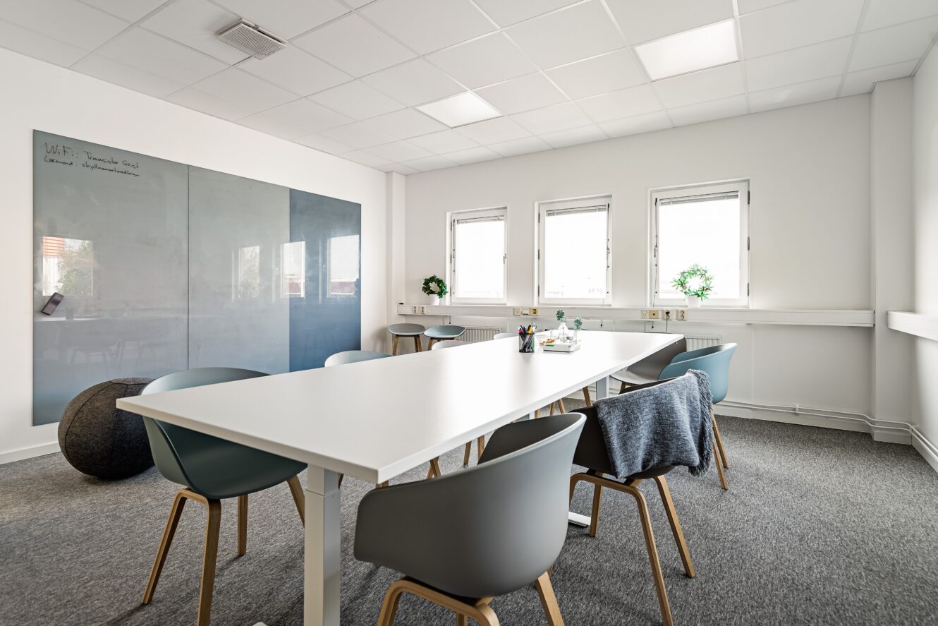 Modernt mötesrum med ett vitt bord, sex stolar, en stor väggmonterad glastavla, tre fönster och minimal inredning i en ljus och ren miljö.