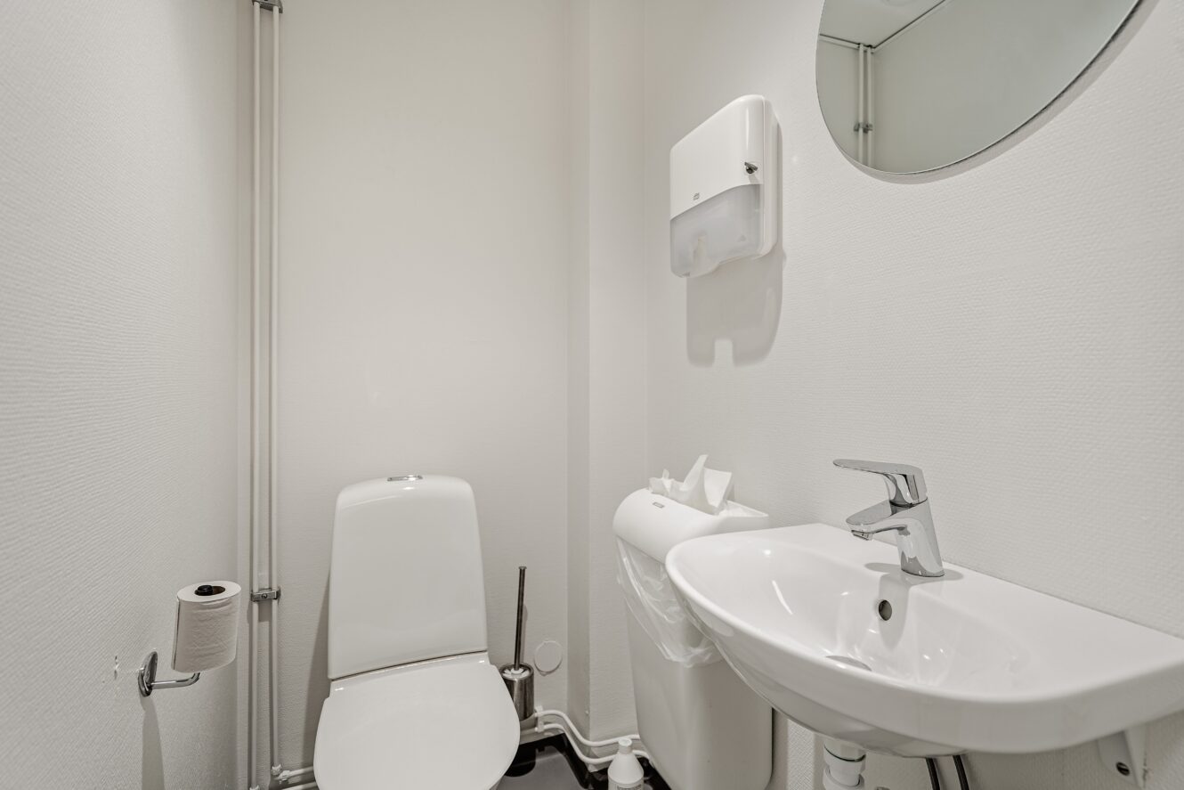 Ett litet badrum med toalett, väggmonterat handfat, rund spegel, toalettpappershållare och en tvålpump monterad ovanför papperskorgen.