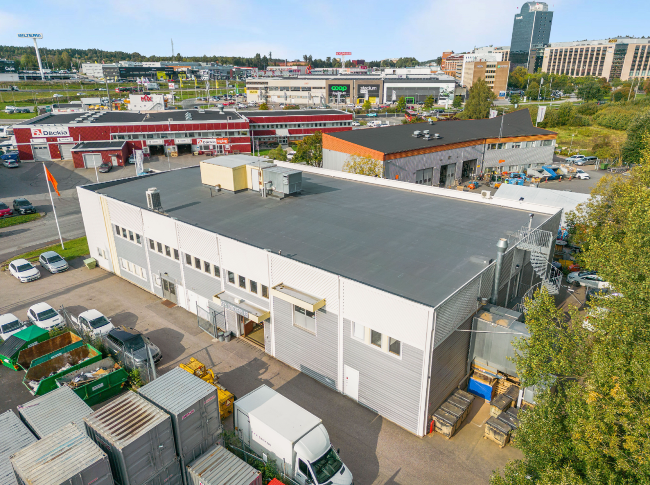 Flygfoto över en stor industrilagerbyggnad med platt tak, omgiven av parkerade fordon, containrar och närliggande kommersiella byggnader.