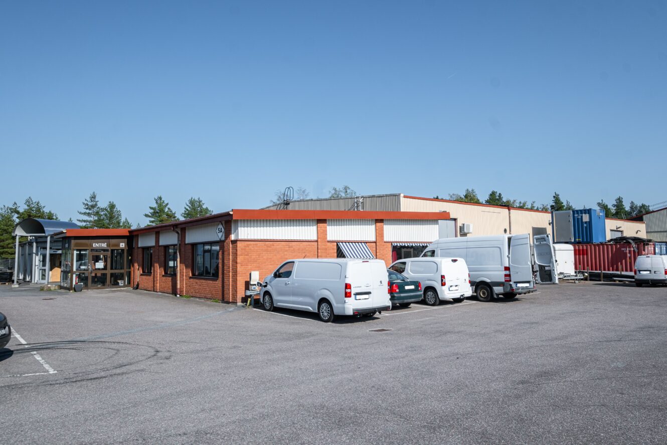 En envånings industribyggnad med en fasad av rött tegel och flera vita skåpbilar parkerade på den intilliggande tomten under en klarblå himmel.