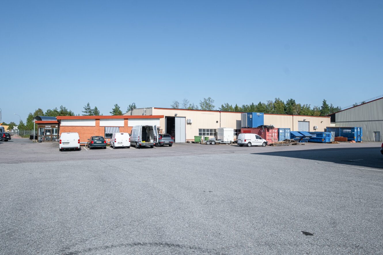 En parkeringsplats med flera vita skåpbilar framför en industriell lagerbyggnad en klar dag. Blå containrar och pallar är staplade i närheten.