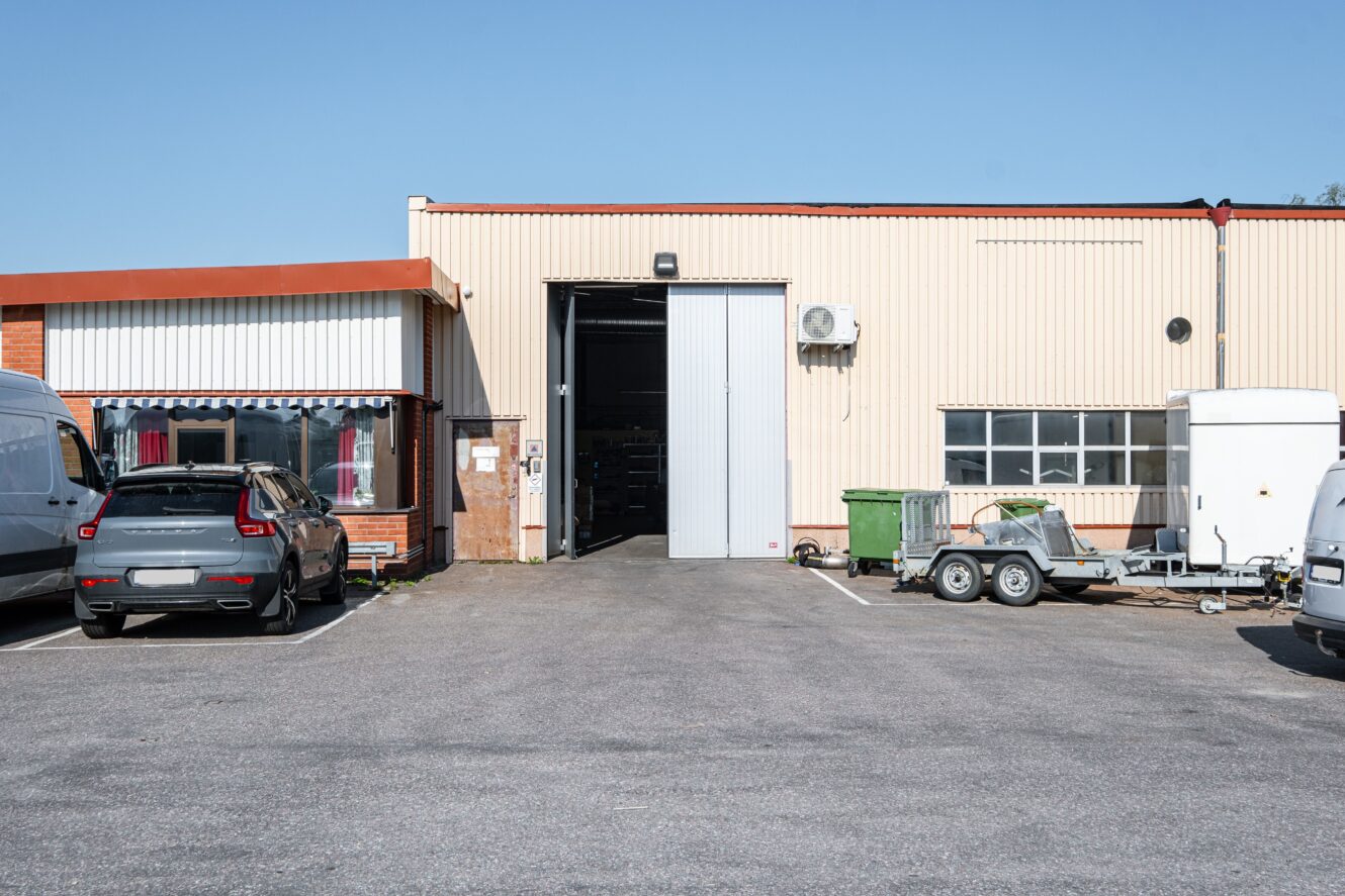 Industriell lagerbyggnad med beige metallfasad, en stor öppen garageport, parkerade bilar, en grön container och en liten släpvagn på parkeringen under en klarblå himmel.