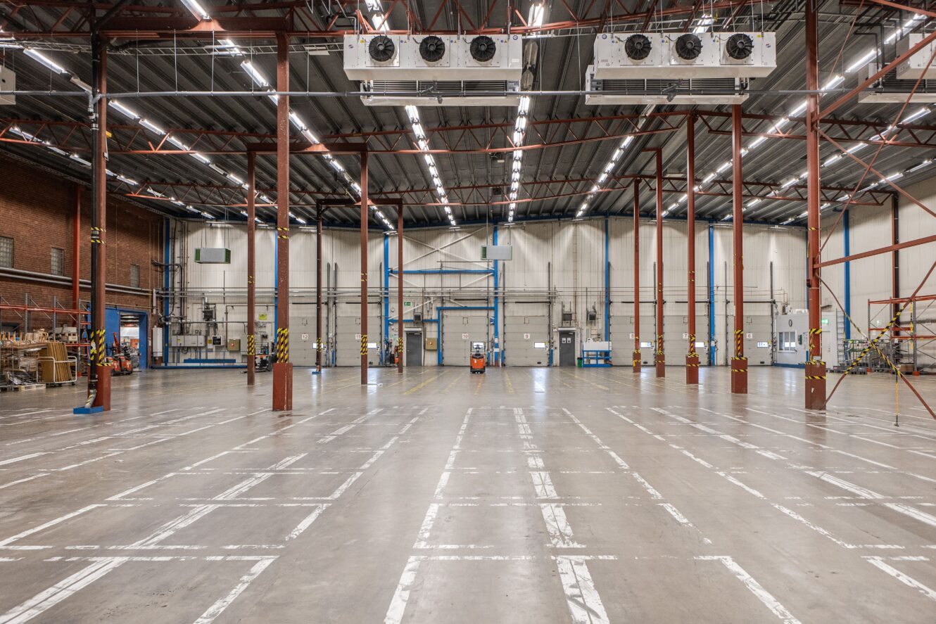 Tomt industrilager med högt i tak, synliga takbjälkar, takbelysning, ventilationsaggregat och markerade linjer på betonggolvet. Hyllor och utrustning syns längs sidorna.