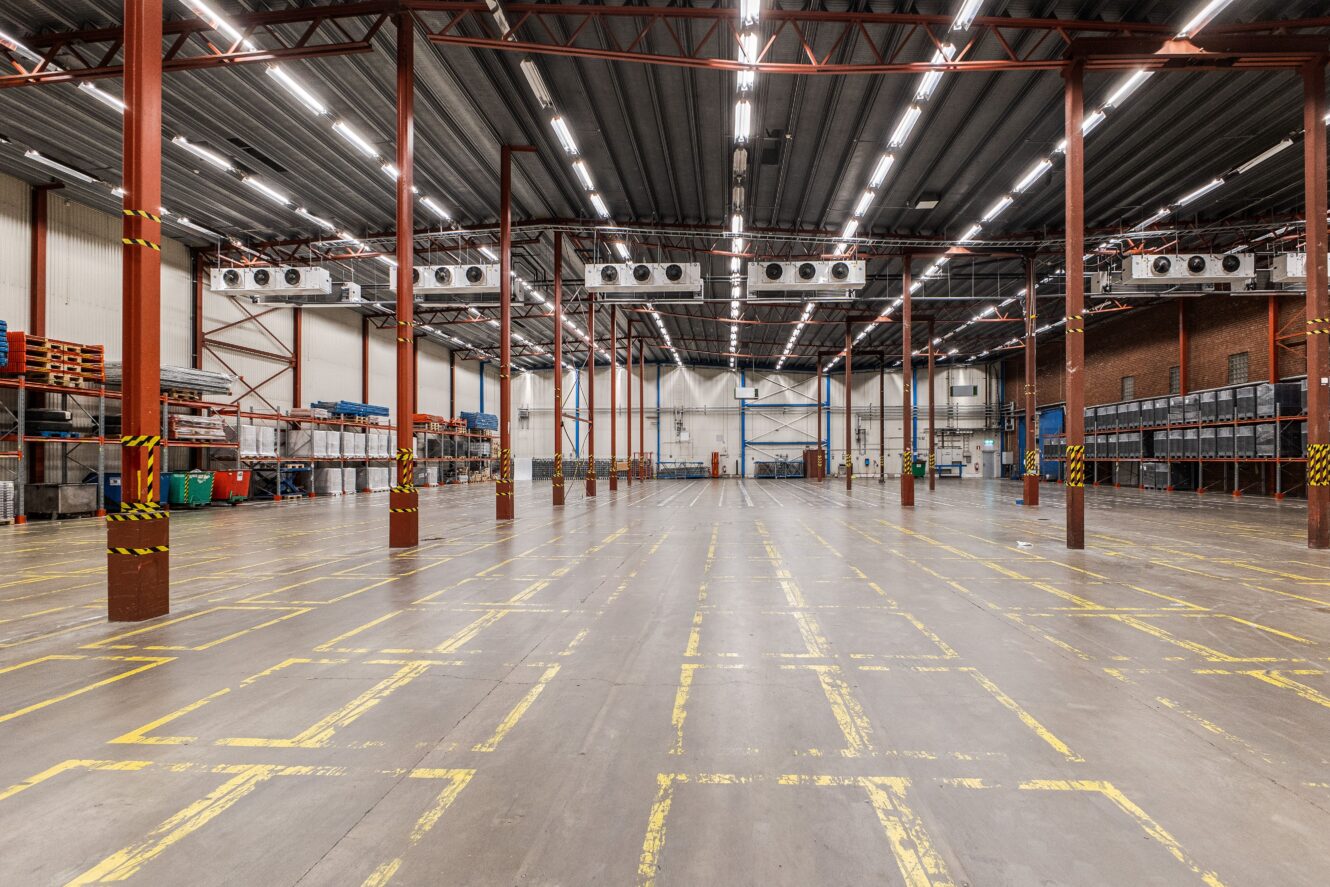 Ett stort, tomt lagerutrymme med höga röda stödbjälkar, takbelysning och markerade ytor på betonggolvet. Hyllor och förvaringslådor står uppställda längs väggarna.