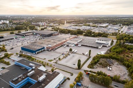 Flygfoto över ett industriområde med stora lagerlokaler, parkerade lastbilar och intilliggande vägar, omgivet av träd och avlägsna bostadshus under en molnig himmel.