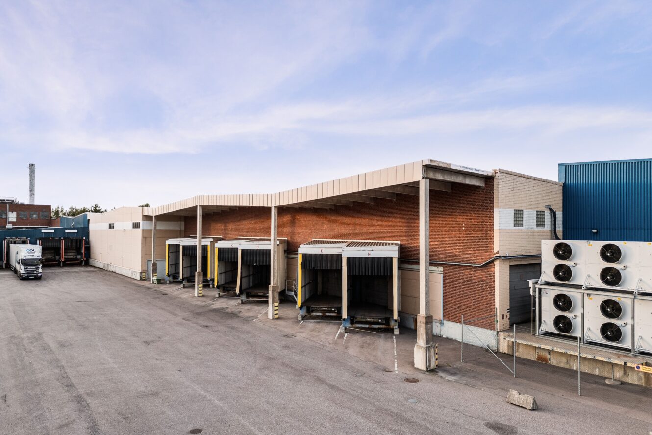 Fyra lastbryggor vid ett kommersiellt lager med en parkerad lastbil i närheten; stora industriella kylaggregat syns till höger i bilden.