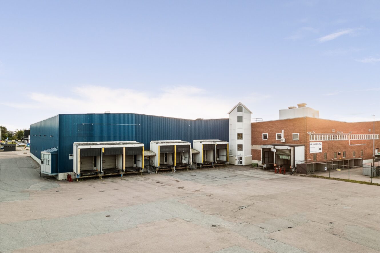 Stor industribyggnad med flera lastkajer, en blå lagerdel, en röd tegeldel och en tom asfalterad tomt framför under en klar himmel.