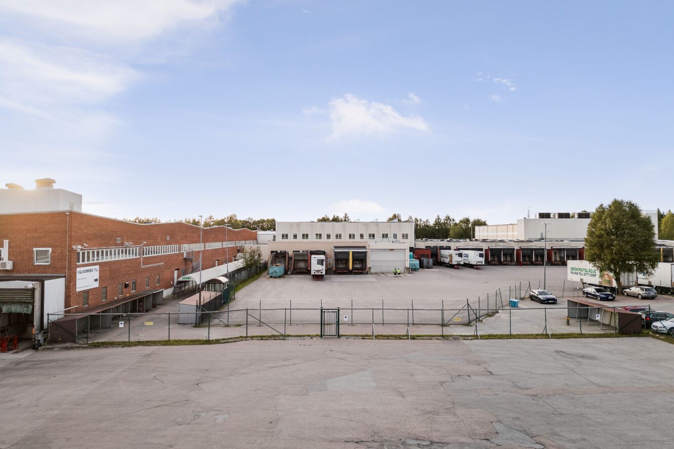 Industrianläggning med flera byggnader, lastkajer, parkerade lastbilar och bilar, omgiven av ett inhägnat område under en delvis molnig himmel.