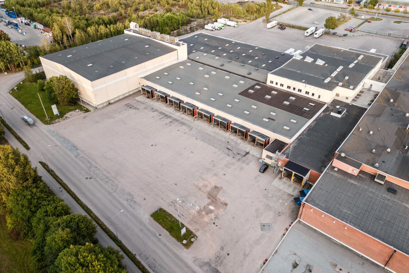 Flygfoto över ett stort industrilager med flera lastkajer, intilliggande parkeringsplatser samt omgivande vägar och grönska.