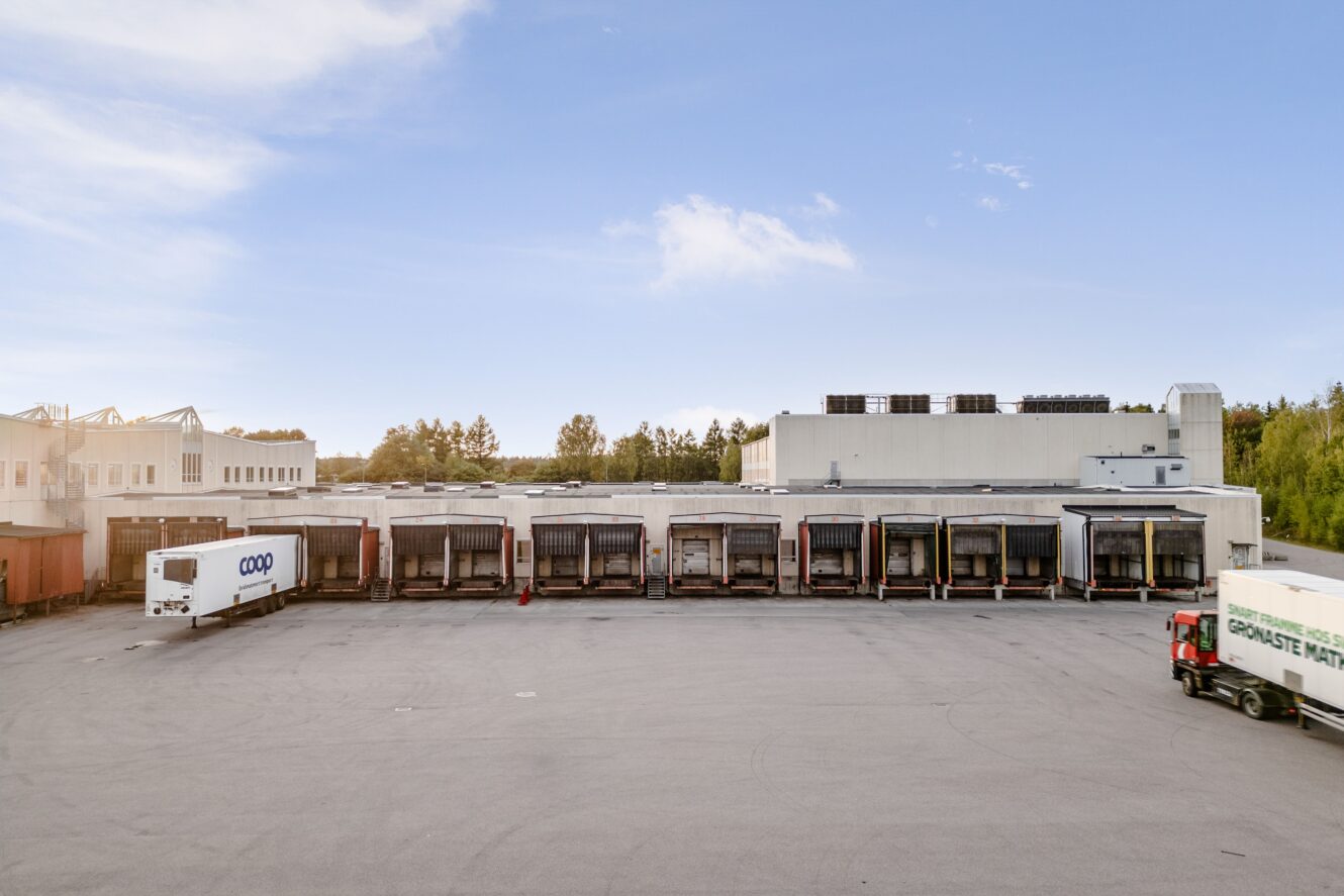 En lagerlokal med flera lastkajer, två vita semitrailers parkerade i närheten och träd i bakgrunden under en klar himmel.
