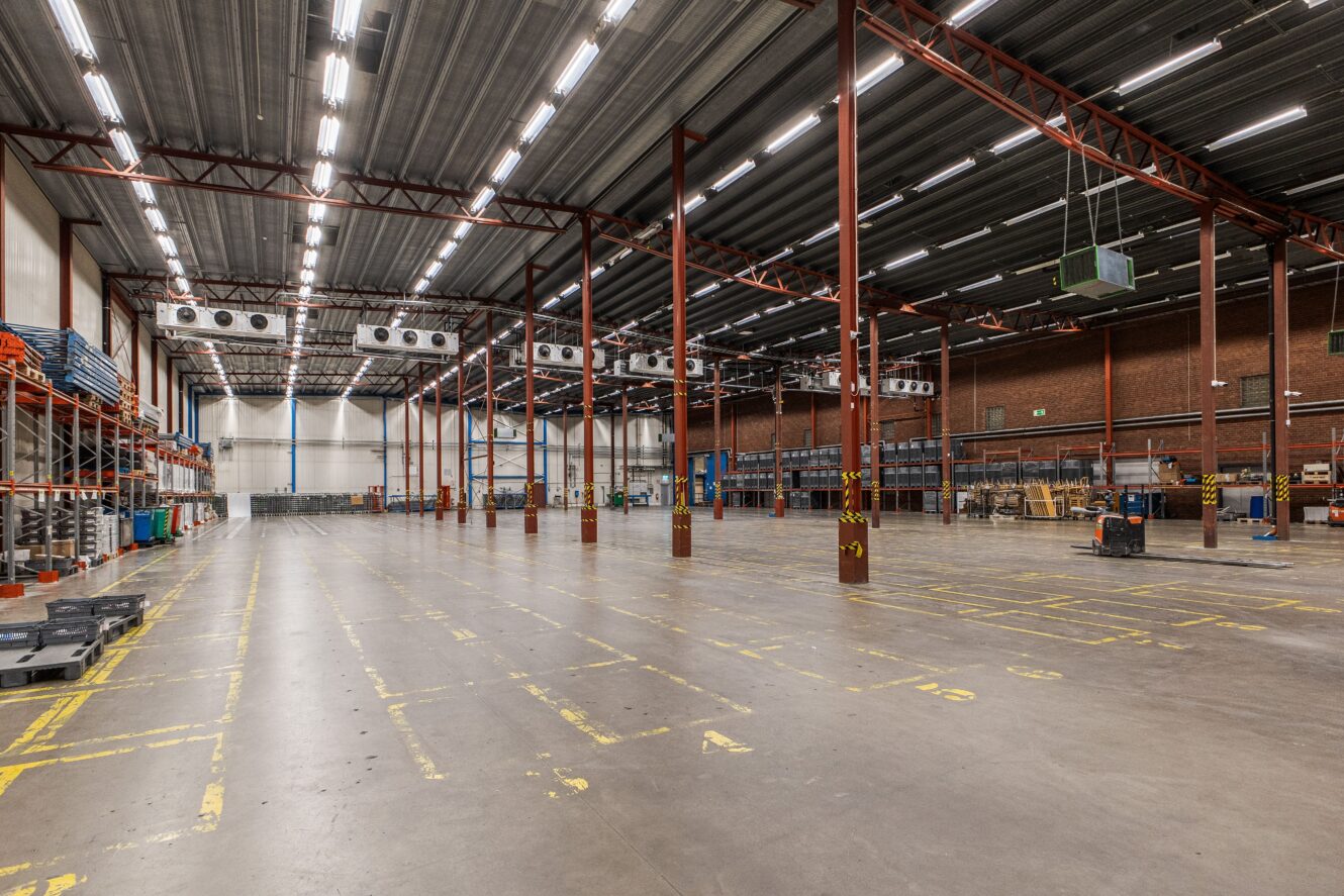 Stor tom lagerlokal med högt i tak, synliga bjälkar, industriell belysning och markerade ytor på det polerade betonggolvet. Hyllor och utrustning står längs hela ytan.