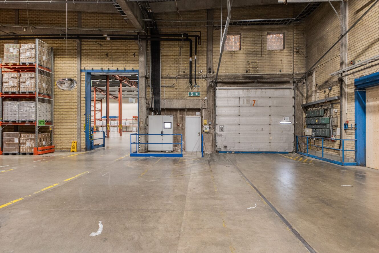 Interiör av en rymlig lagerlokal med betonggolv, tegelväggar, hyllor fyllda med lådor, en stor garageport och synliga elpaneler.