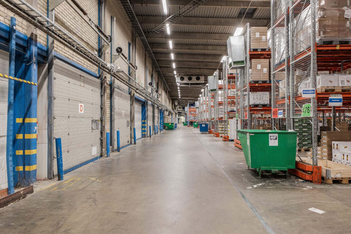 Bred interiörvy av ett lager med höga hyllor som lagrar lådor och gröna behållare längs en central betonggång.