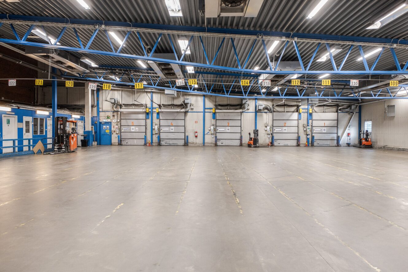 Stor, tom lagerlokal med en rad numrerade lastkajdörrar, högt i tak och industriell belysning.