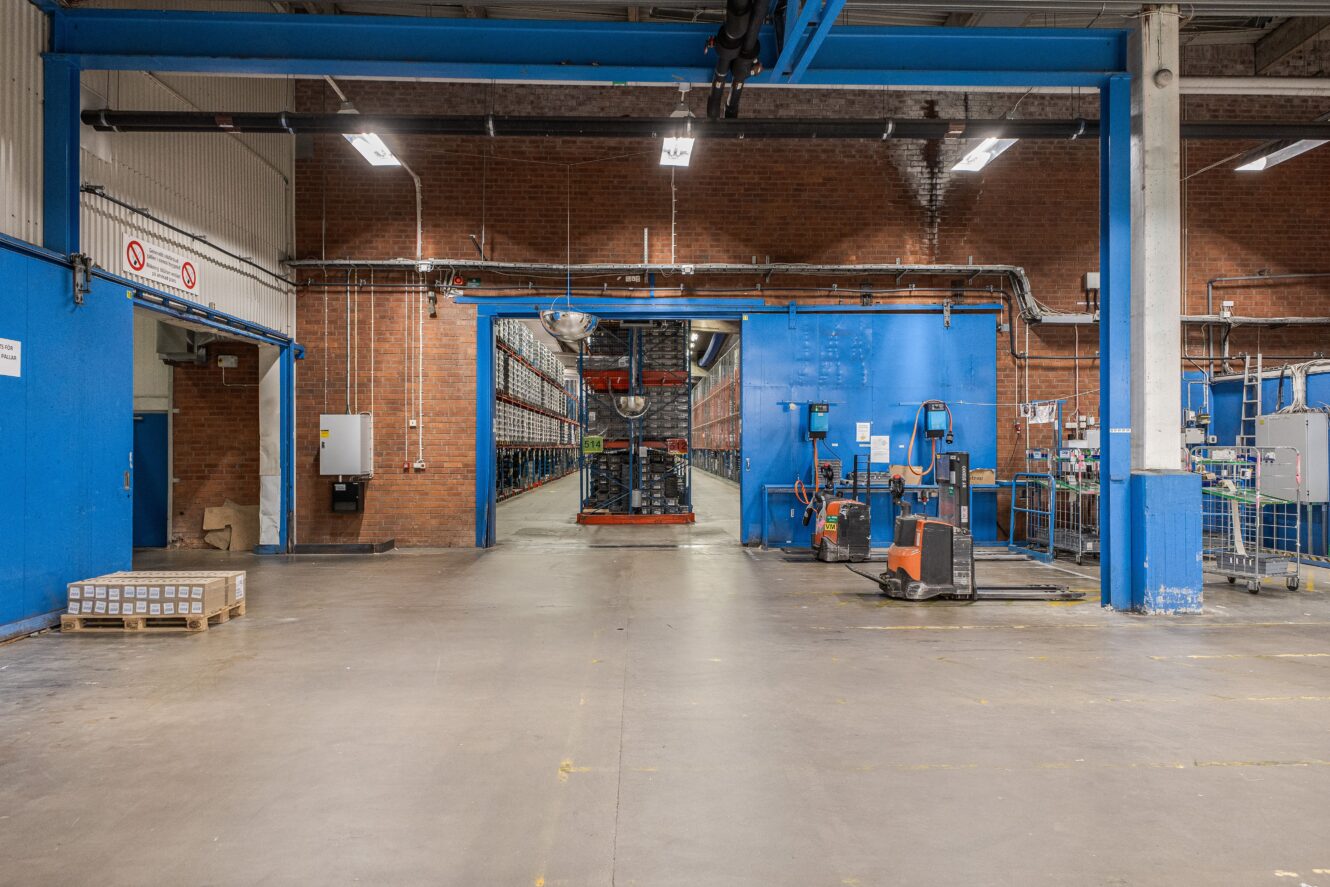 En lagerlokal med blå dörrar, industrihyllor fyllda med varor och två orange elektriska pallbockar nära ingången.
