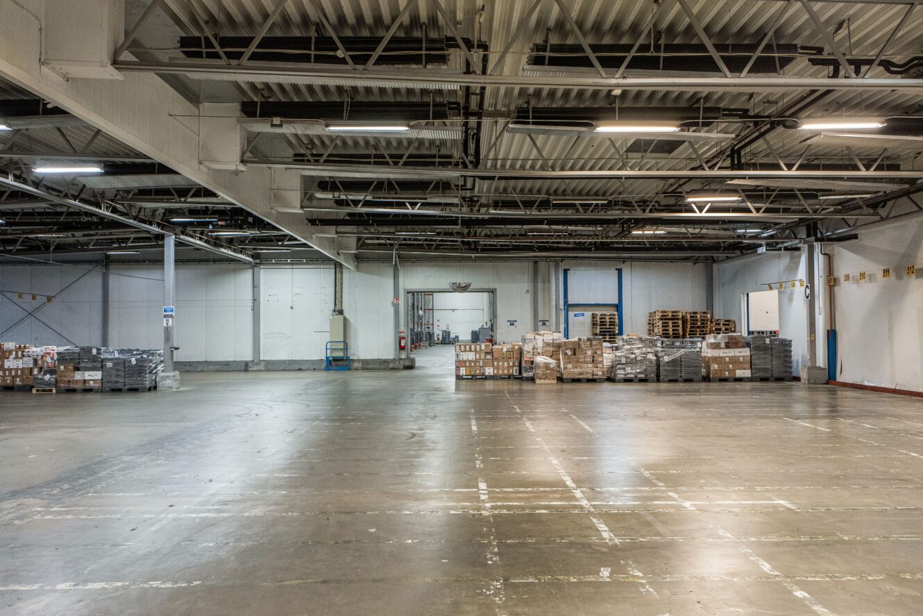 Stor, tom lagerlokal med några staplar med pallar och lådor längs bakväggen, synliga takbjälkar och polerat betonggolv.