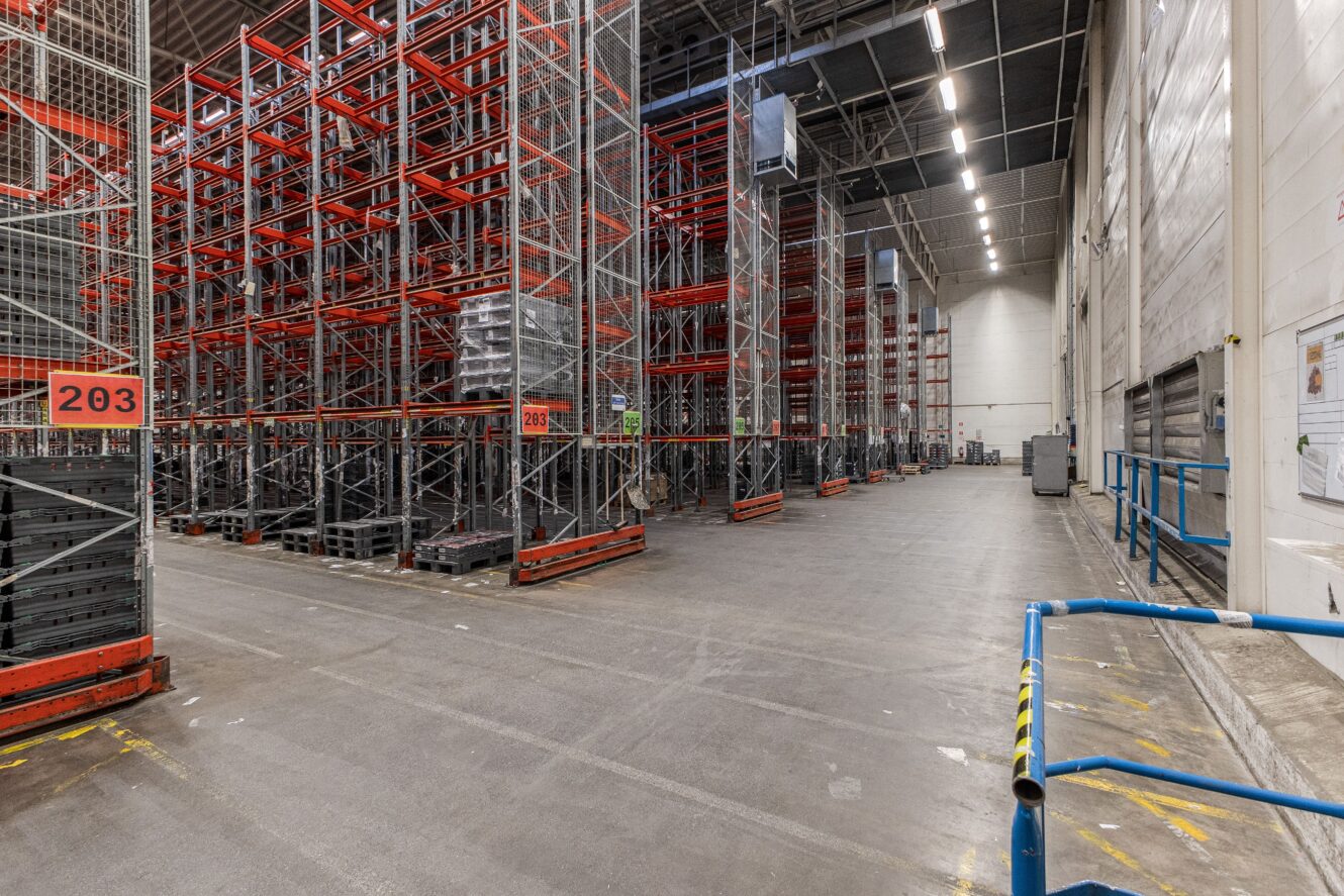 Stor lagerlokal med höga lagerhyllor i metall, pallade varor, betonggolv och stark takbelysning. En orange skylt med texten 203 syns till vänster.
