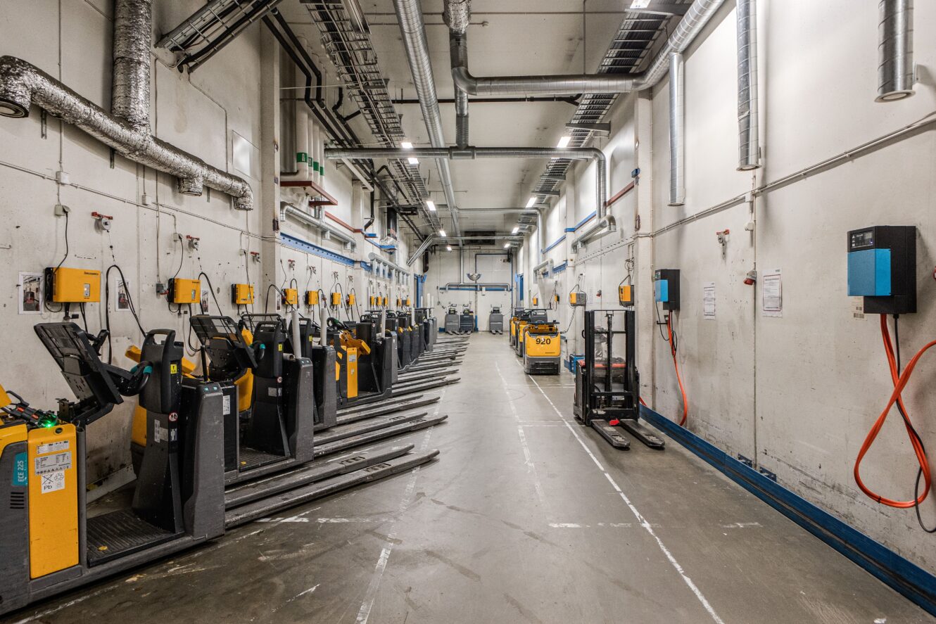Industrihall med flera pallvagnar dockade på båda sidor, laddningsutrustning monterad på väggarna och synliga rör och kablar ovanför.