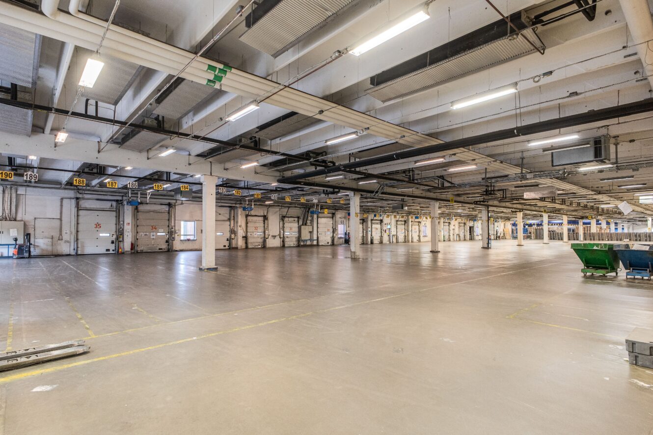 Stort, tomt industrilager med betonggolv, högt i tak, takbelysning, stödpelare och flera lastbryggor längs ena väggen.