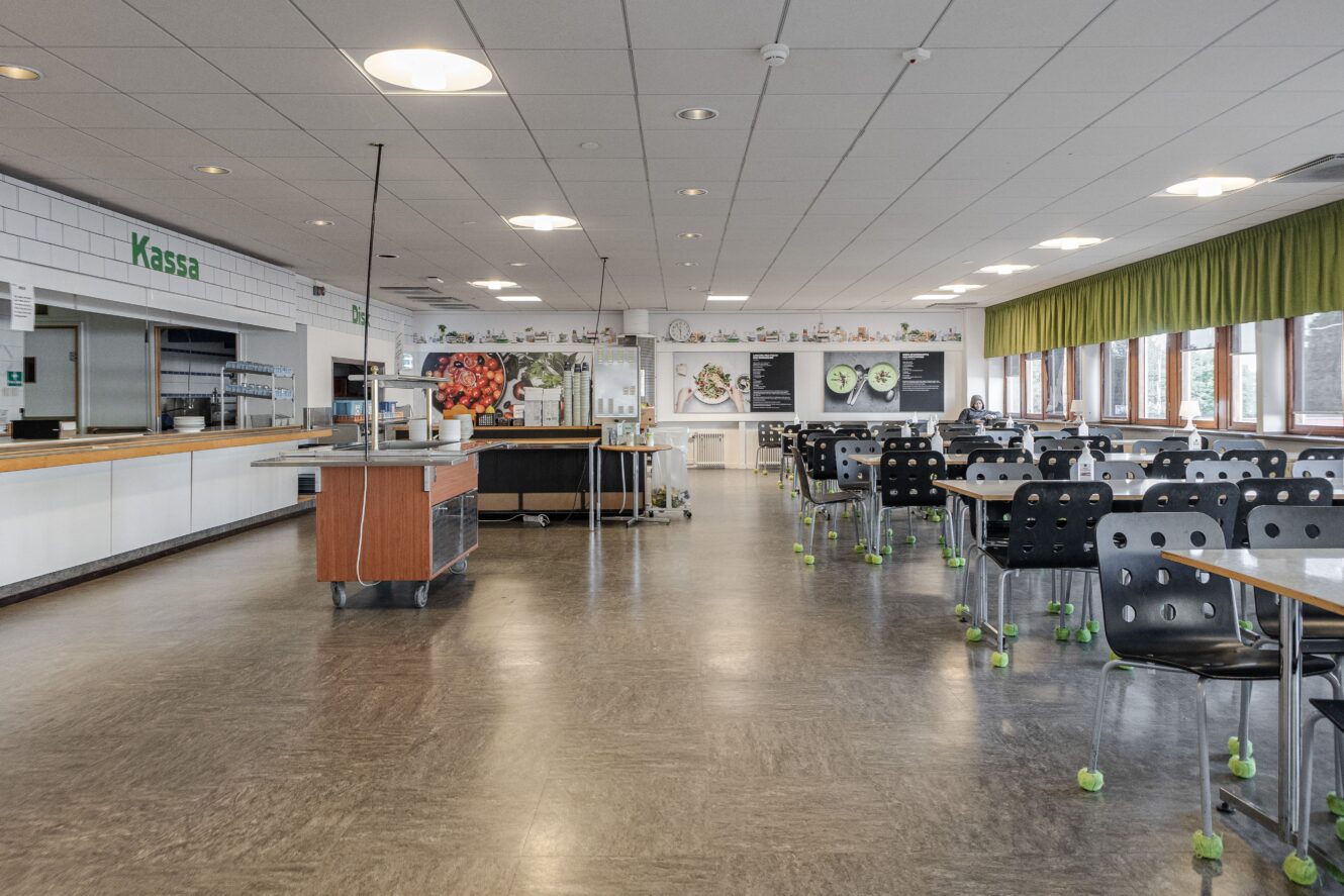 En rymlig, tom cafeteria med svarta stolar, ljusa bord, en serveringsdisk och stora fönster med gröna gardiner längs ena väggen.