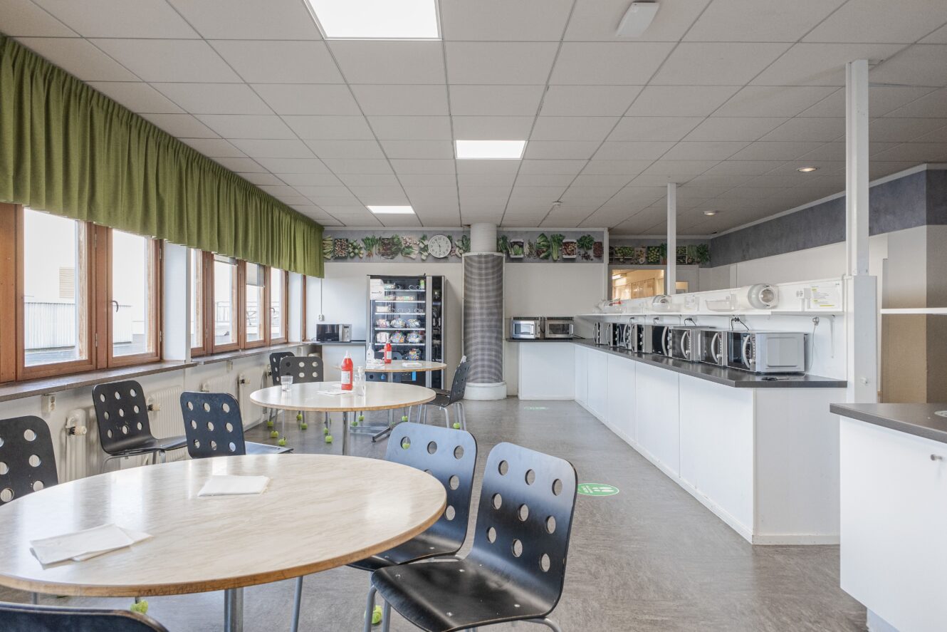 Ett rent, modernt fikarum med runda bord, svarta stolar, mikrovågsugnar, en varuautomat och stora fönster med gröna gardiner.