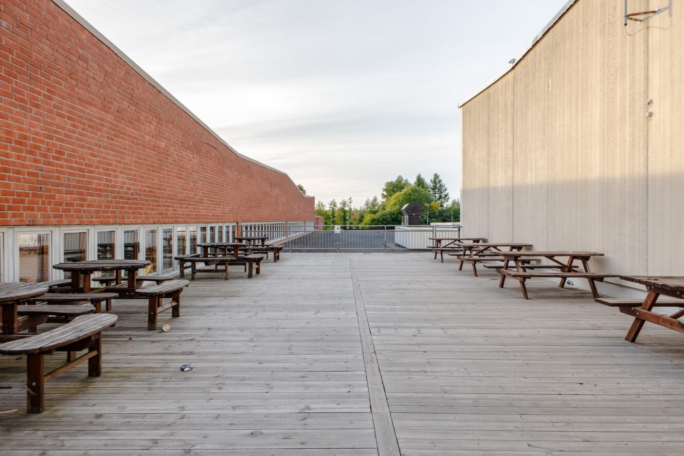 Trädäck med flera picknickbord placerade mellan två byggnader, en i tegel och en i betong, med träd synliga i bakgrunden.