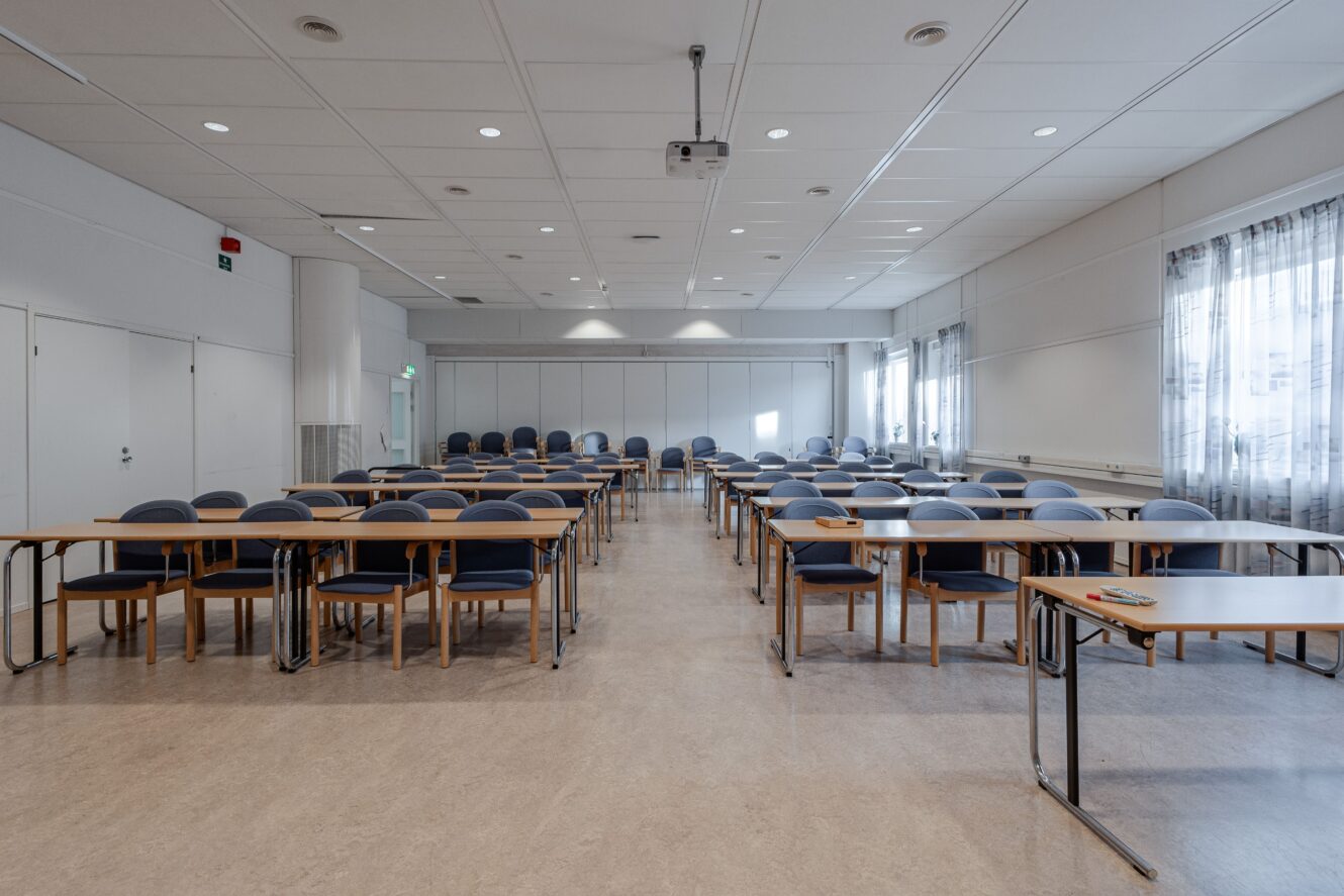 Ett tomt klassrum med rader av bord och stolar som vetter mot en projektorduk, upplyst av taklampor och naturligt ljus från fönster med skira gardiner.