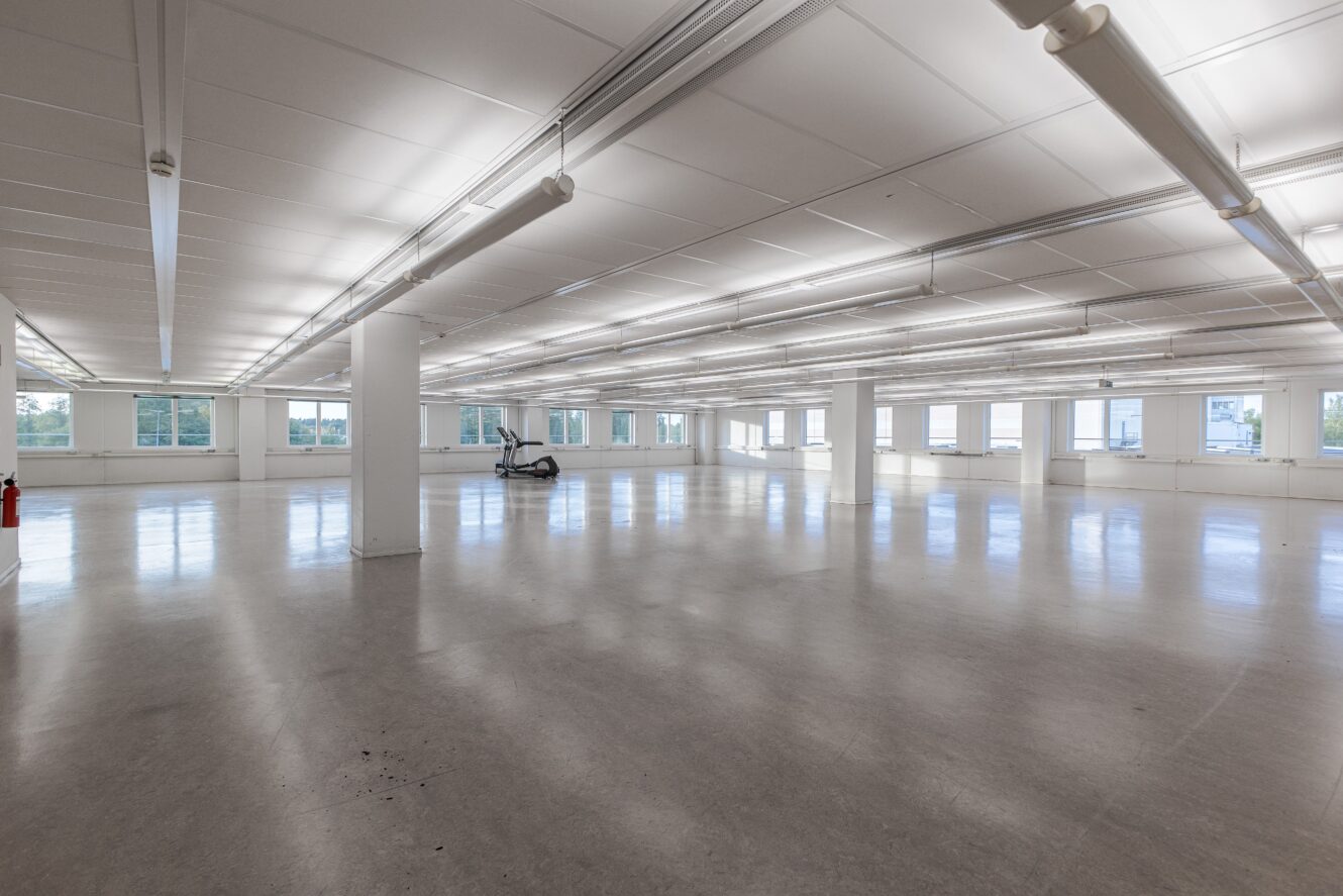 Stort tomt kontorsutrymme med vita väggar, taklampor, många fönster och en enda motionscykel nära den bakre väggen.