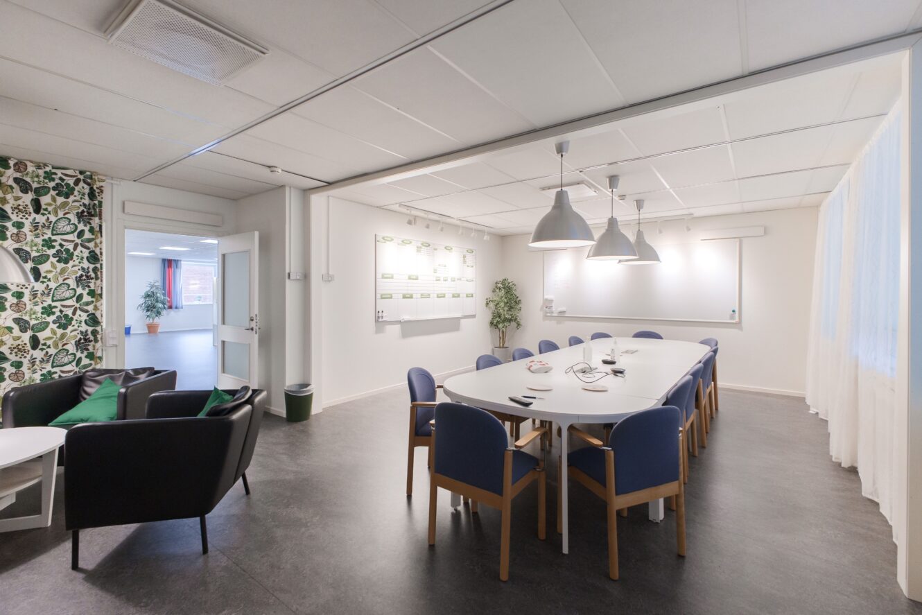 Ett modernt konferensrum med ett långt vitt bord, blå stolar, pendellampor, whiteboardtavlor och en sittgrupp med fåtöljer och mönstrad tapet.