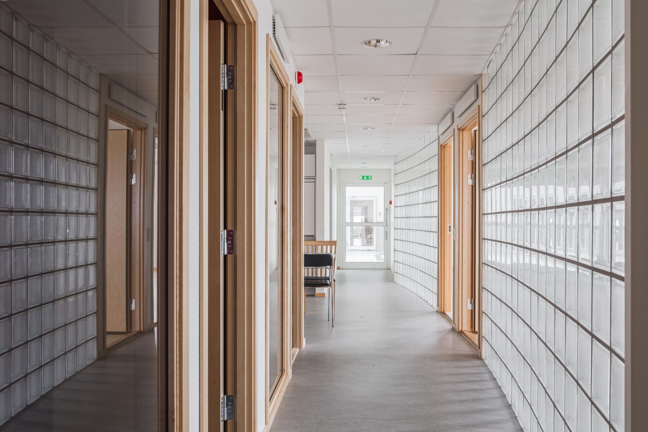 En modern hall med väggar av glasblock, dörrkarmar av trä, grått golv och en stol nära en utgång i slutet, upplyst av naturligt ljus.